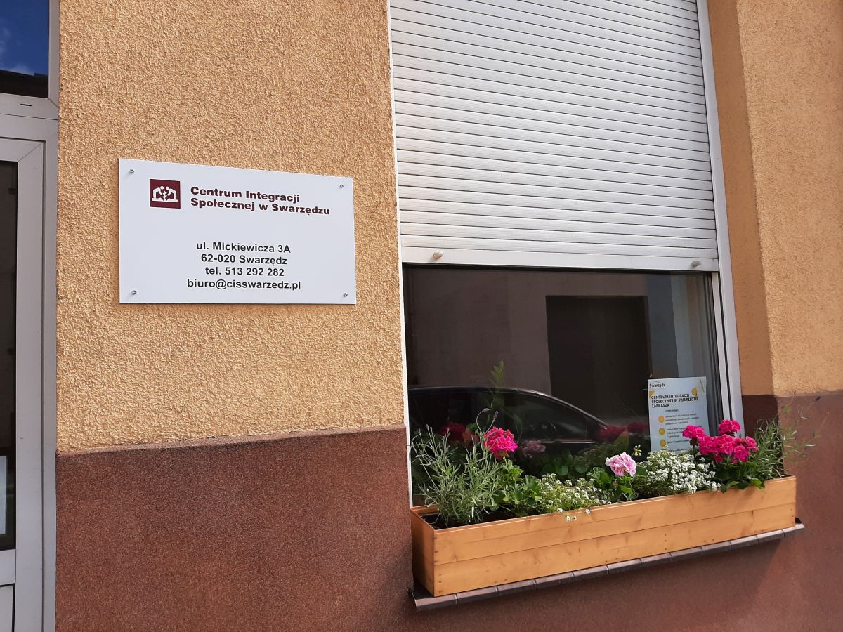Tablica informacyjna na budynku Centrum Integracji Społecznej w Swarzędzu z adresem, numerem telefonu i adresem e-mail. Obok okno z zamkniętą roletą i skrzynka z kwiatami
