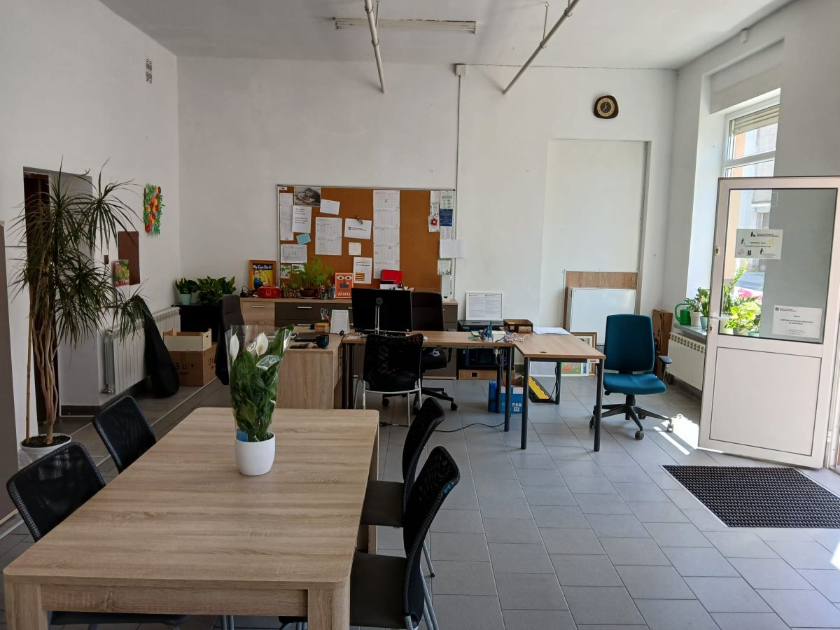 Biuro Centrum Integracji Społecznej z biurkami, tablicą informacyjną, roślinami i stołem dla interesantów. W tle otwarte drzwi i okno z kwiatami na parapecie.