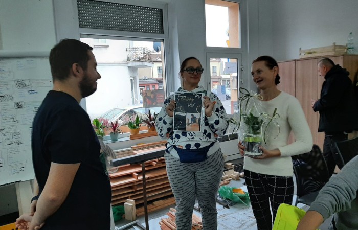 Urodzinowe spotkanie w pomieszczeniu – kobieta trzyma roślinę w doniczce, a druga osoba prezentuje kartkę urodzinową. Na ścianach widać ozdoby oraz notatki.