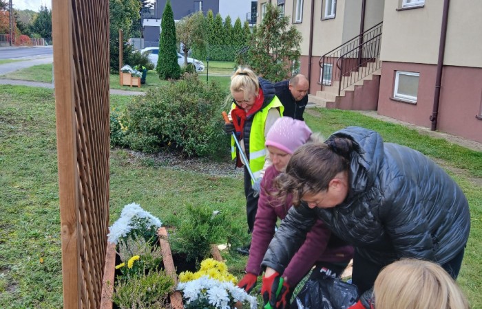 Grupa osób wspólnie sadzi jesienne kwiaty w drewnianej donicy przed budynkiem mieszkalnym. Obok donicy widoczna jest kratka pergoli.