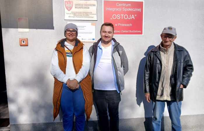  Trzy osoby stojące przed budynkiem Centrum Integracji Społecznej „Ostoja” w Pniewach, pozujące do zdjęcia przy tablicy informacyjnej.