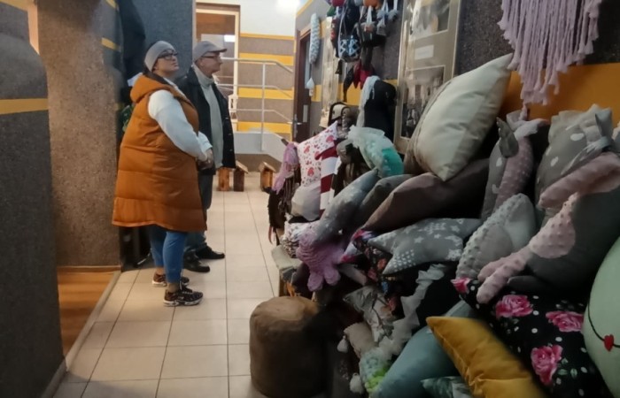 Dwie osoby oglądające ręcznie wykonane poduszki i maskotki, wyeksponowane na ścianach korytarza budynku.