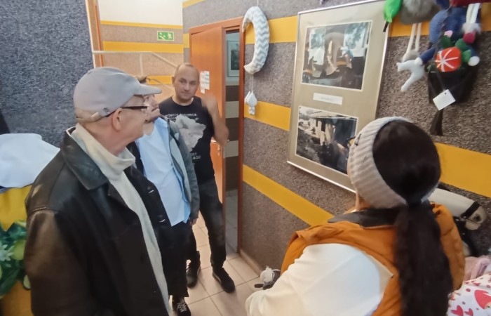 Grupa uczestników oglądająca zdjęcia i pamiątki na ścianie korytarza, prowadzona przez przewodnika opowiadającego o eksponatach.